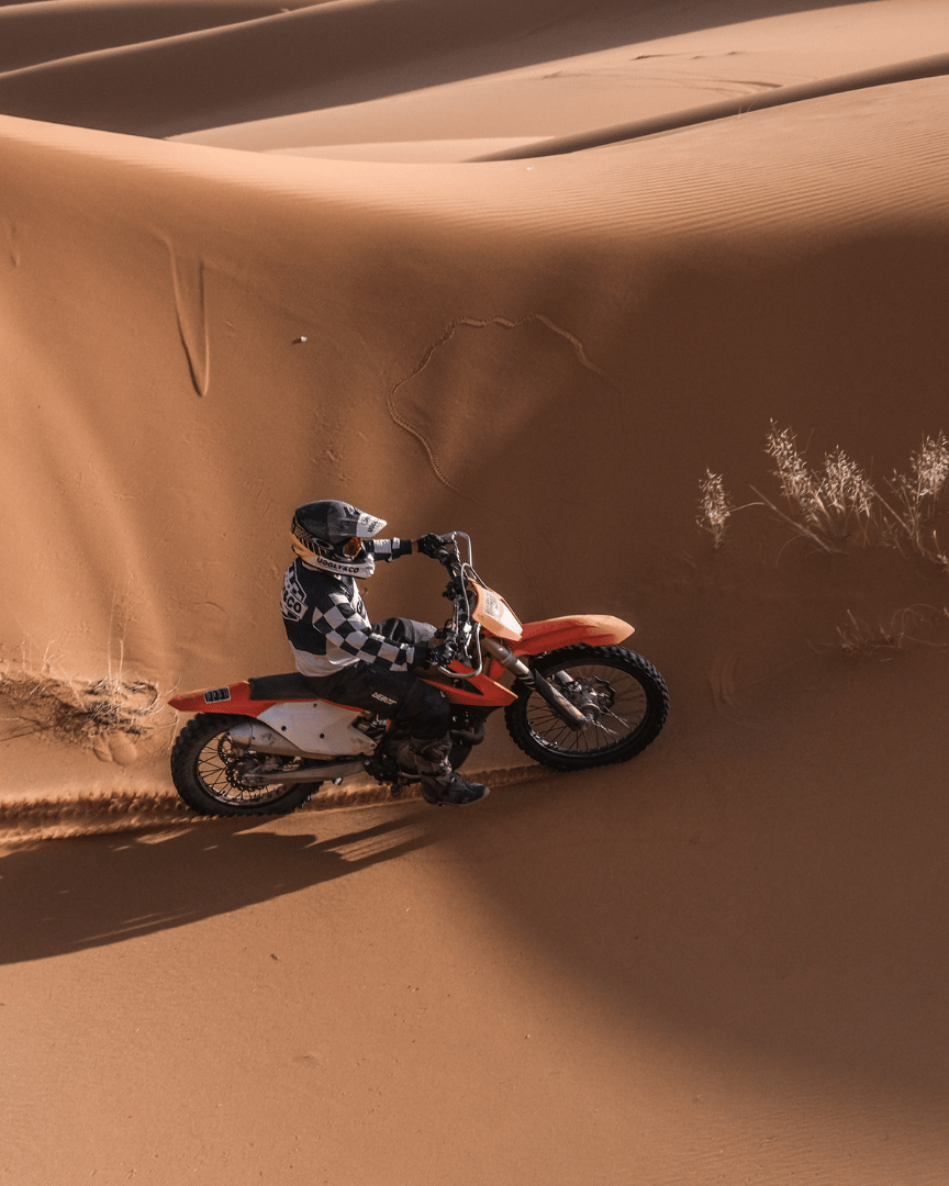 A person riding a motocross bike across sand dunes in full gear in the desert wearing a chequered Uggly&Co MX shirt