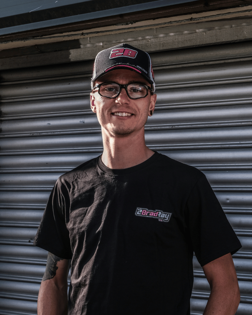 Front view of Bradley Ray wearing a trucker hat with number 28 and a black t-shirt with branding, standing against a garage roller door