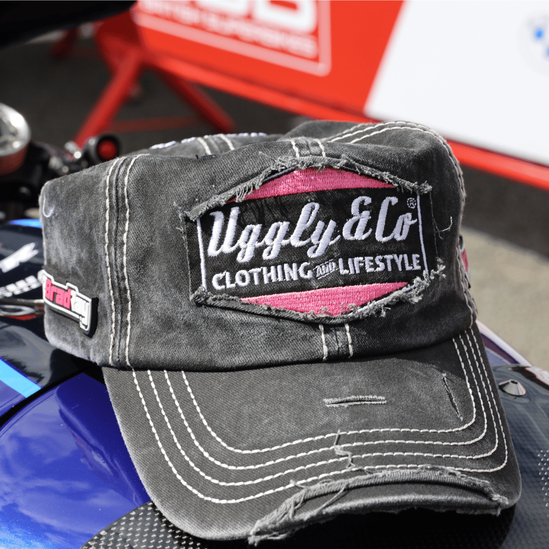 A black trucker hat with the Uggly&Co logo in silver and pink on the front, and additional Bradley Ray branding on the side, resting on a motorcycle fuel tank