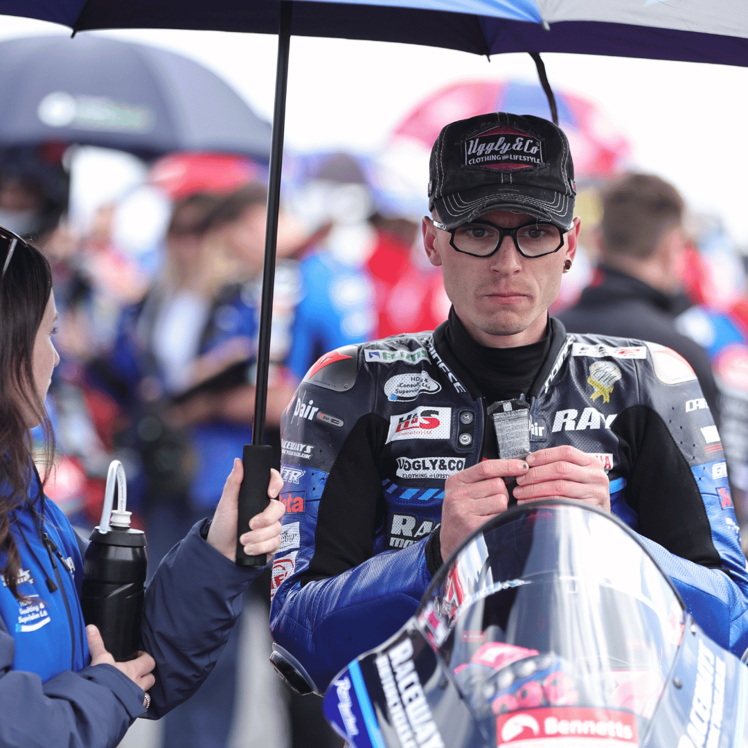Bradley Ray wearing a black trucker hat with the Uggly&Co logo in silver and pink on the front, in the racing paddock on his motorcycle.