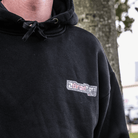 Black hoodie with a logo on the chest worn by a person, outdoors.
