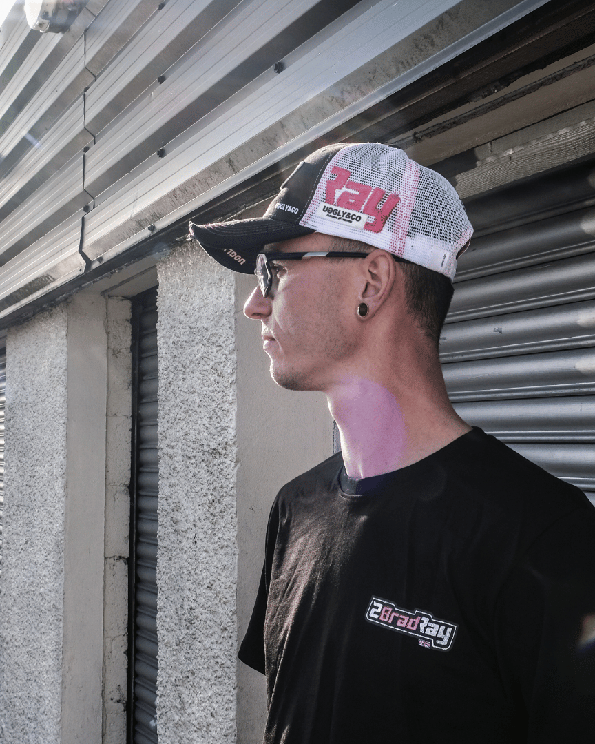 Side view of Bradley Ray wearing a trucker hat with number 28 and a black t-shirt with branding, standing against a garage roller door
