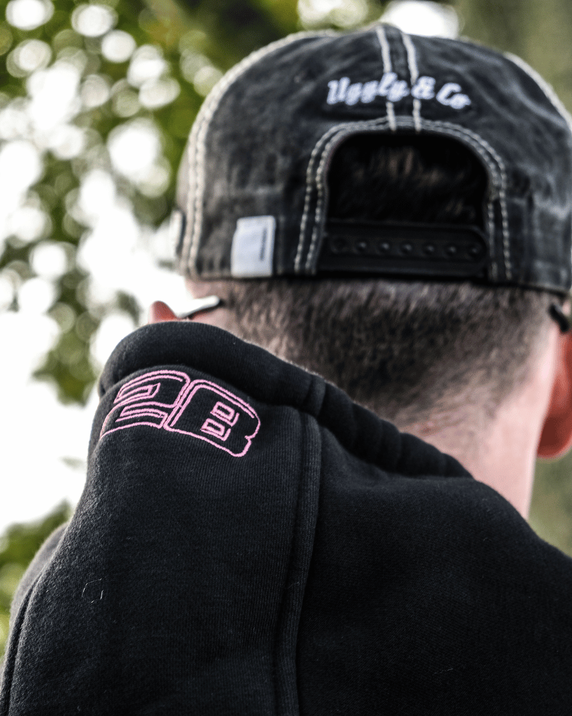 Person wearing a black cap and black hoodie with pink logo, standing outdoors.