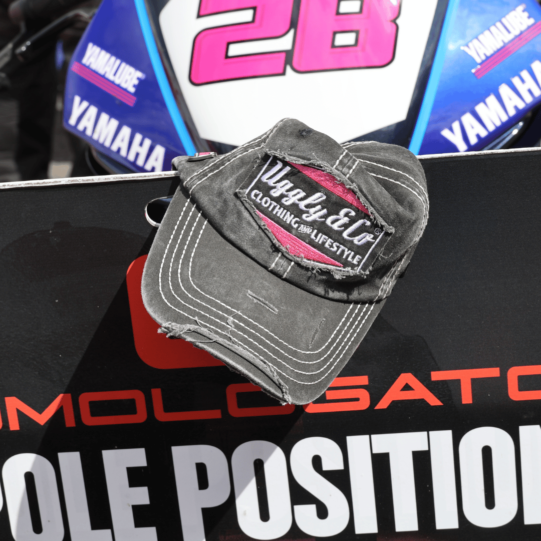 A black trucker hat with the Uggly&Co logo in silver and pink on the front resting on a sign in front of a motorcycle