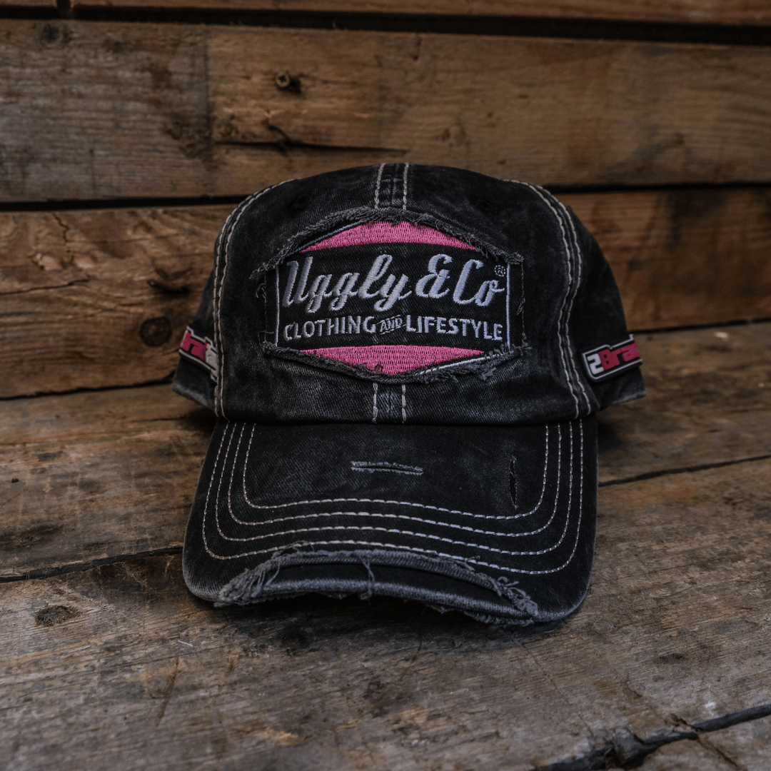 Front view of a black trucker hat with the Uggly&Co logo in silver and pink on the front, and additional Bradley Ray branding on the side, set against a vintage wooden background.