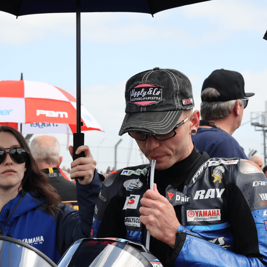 Bradley Ray wearing a black trucker hat with the Uggly&Co logo in silver and pink on the front, and additional Bradley Ray branding on the side, in the racing paddock.