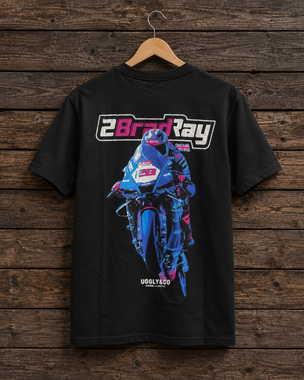A black short-sleeve t-shirt hanging on a wooden hanger against a rustic dark wood plank wall. The back of the shirt features a large graphic print of a blue and pink motorcycle racer with the text '2BradRay' above it and 'UGGLY CO' below it.