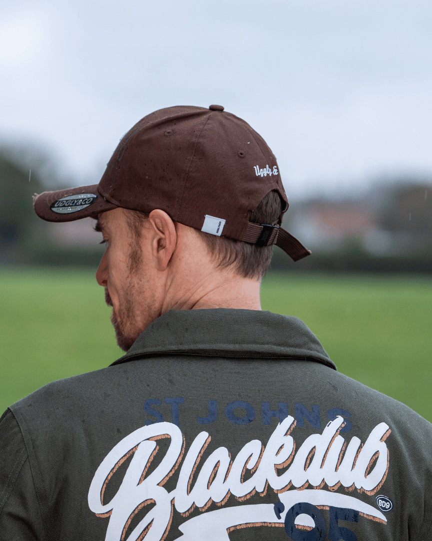 Rear view of a man while wearing a brown jacket and trucker hat with 'Uggly & Co' branding with a blurred field background