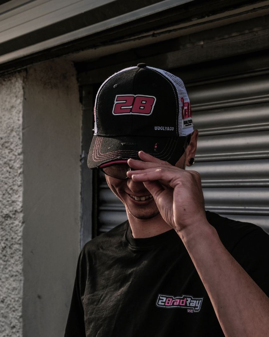 Bradley Ray wearing a trucker hat with number 28 and a black t-shirt with branding, standing against a garage roller door