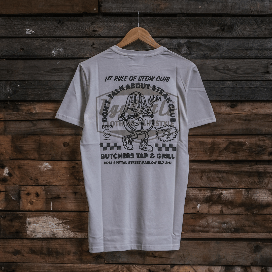Rear view of a Tom Kerridge's Butchers Tap and Grill white t-shirt with black graphics and text on a wooden background
