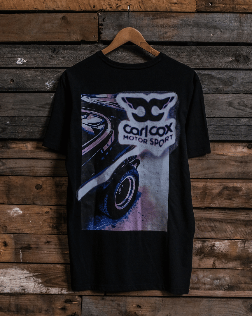 Rear view of a Carl Cox Motorsport black t-shirt with car graphic and 'carl cox motorsport' logo on a wooden background