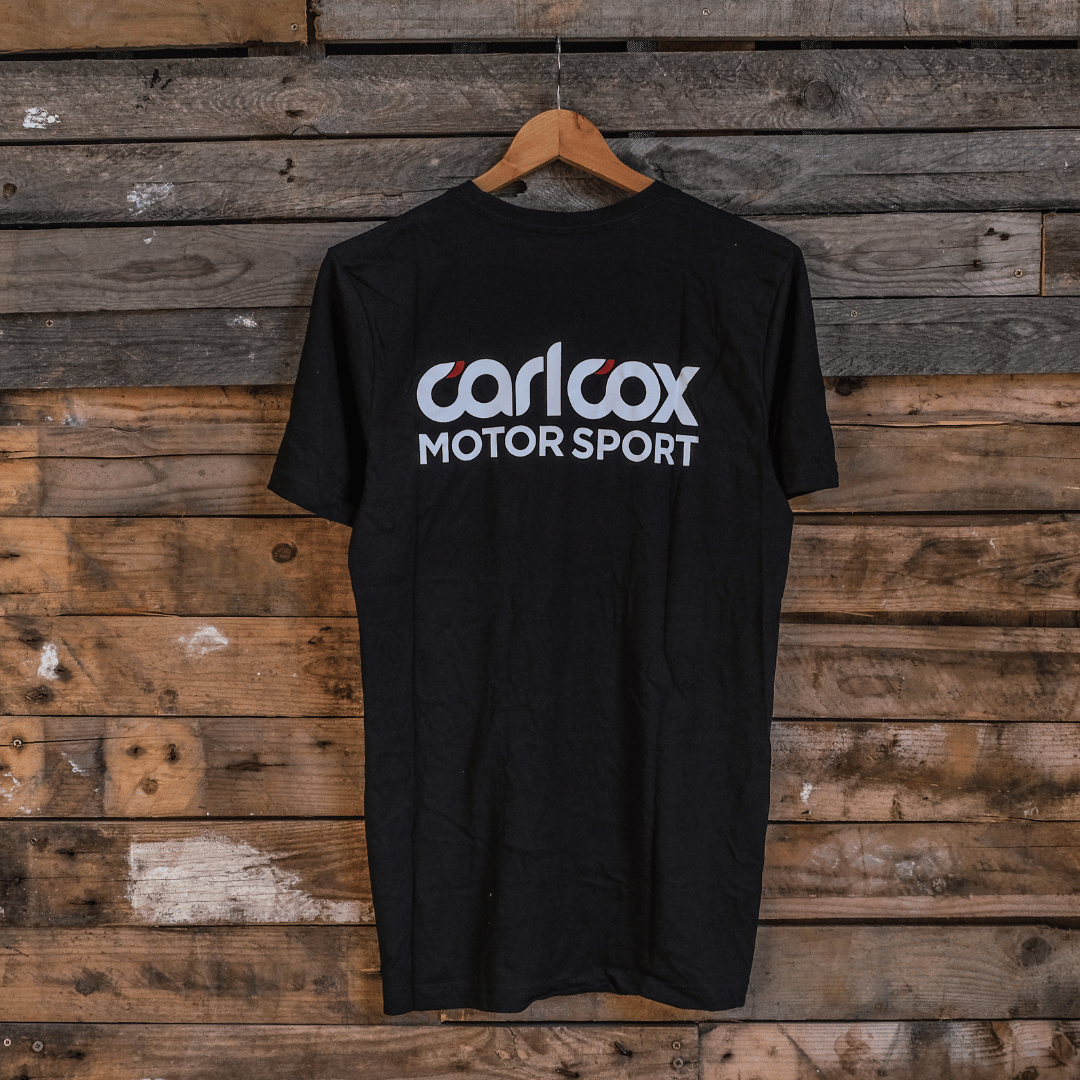 A black t-shirt with 'carl cox motorsport' text printed on the front, displayed against a wooden background.