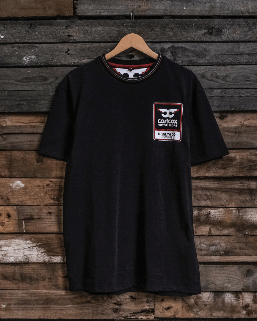 Front view of a Carl Cox motorsport black t-shirt with an Uggly&Co logo on a wooden background