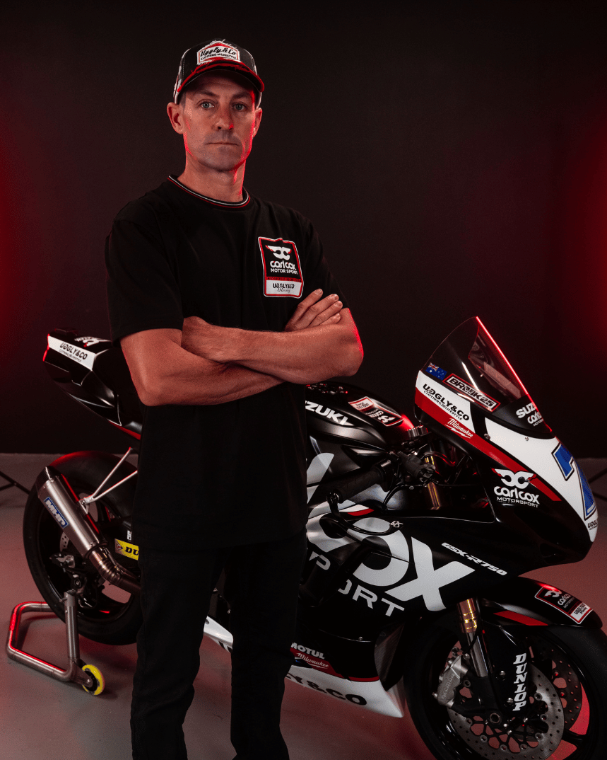 A model wearing a Carl Cox motorsport black t-shirt with an Uggly&Co logo facing the camera with a motorcycle behind