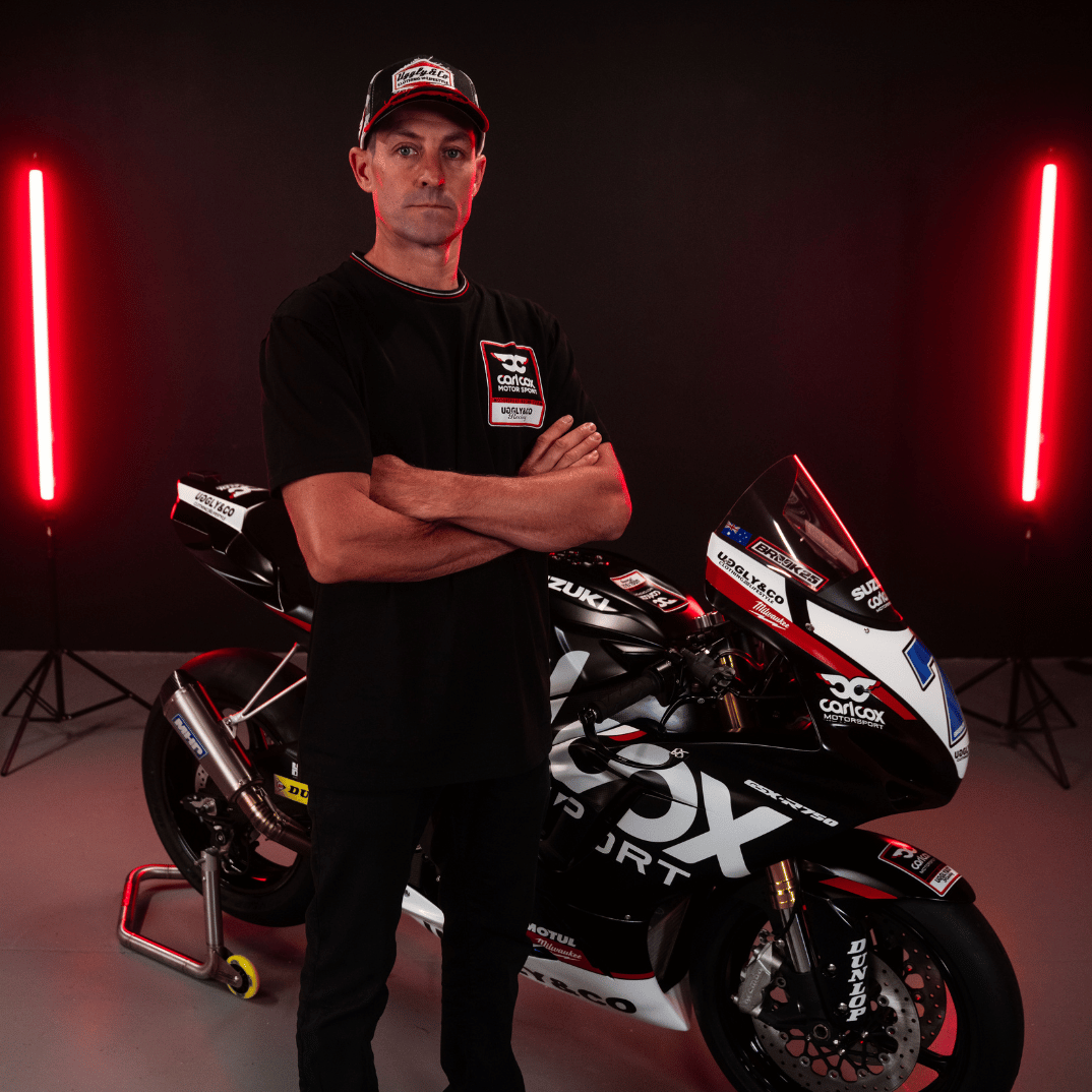 A model wearing a Carl Cox motorsport black t-shirt with an Uggly&Co logo facing the camera with a motorcycle behind