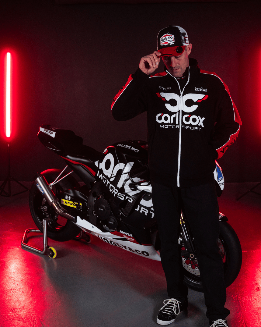 Josh Brookes wearing black and red jacket with Carl Cox Motor Sport, Uggly&Co and Team Classic Suzuki branding in front of a motorbike
