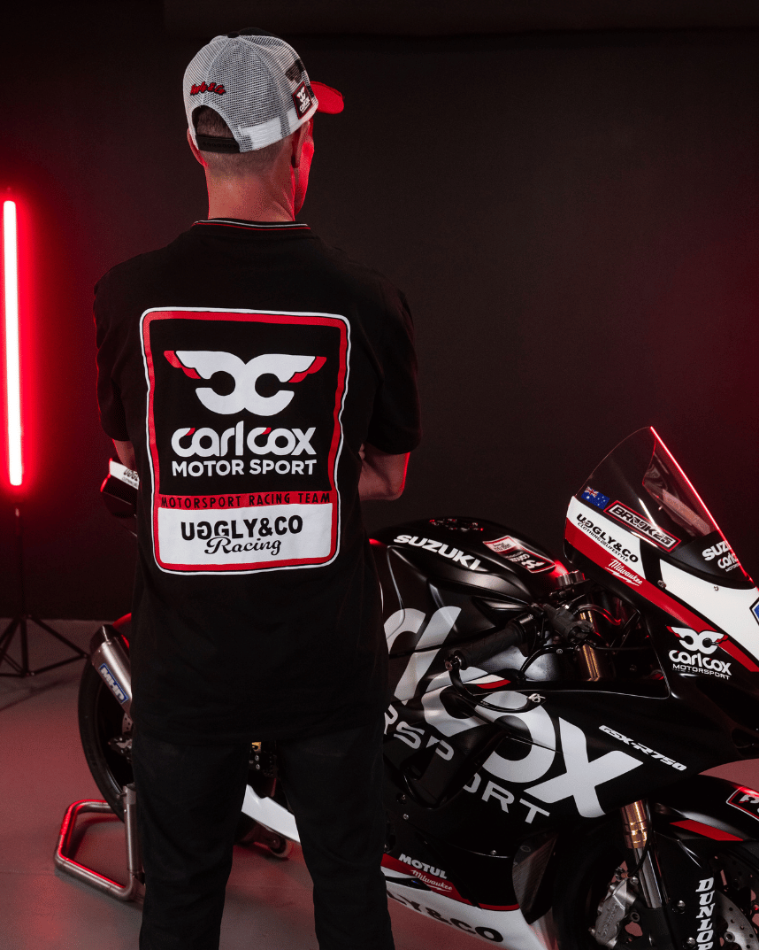 Rear view of a model wearing a Carl Cox motorsport black t-shirt with an Uggly&Co logo with a motorbike in the background