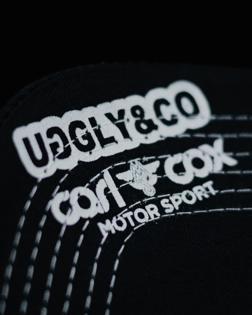 Close up of the Uggly&Co and Carl Cox Motorsport logos on the under side of the peak of an Official Carl Cox Motorsport Merchandise trucker hat in black and white