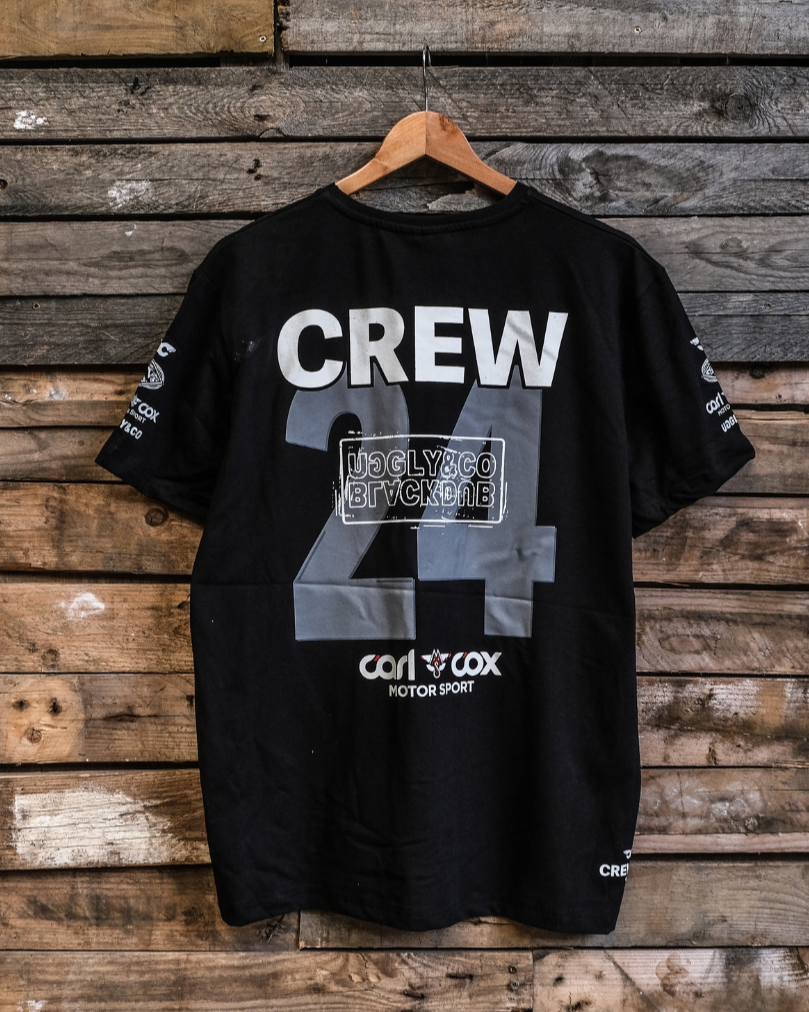 A black crew t-shirt with the text 'CREW 24' and logos printed on the front, displayed on a wooden background.