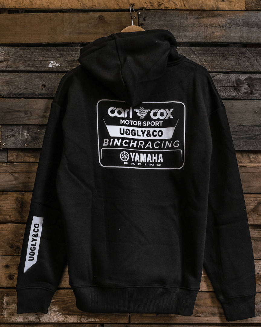 A black hoodie with white and grey text and logos, including 'Carl Cox Motorsport' and 'Uggly&Co', displayed on a wooden background.