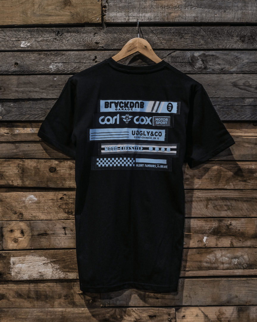 A black crew neck t-shirt with a graphic design in the center, featuring text and a cassette tape motif. The t-shirt is displayed on a wooden background.