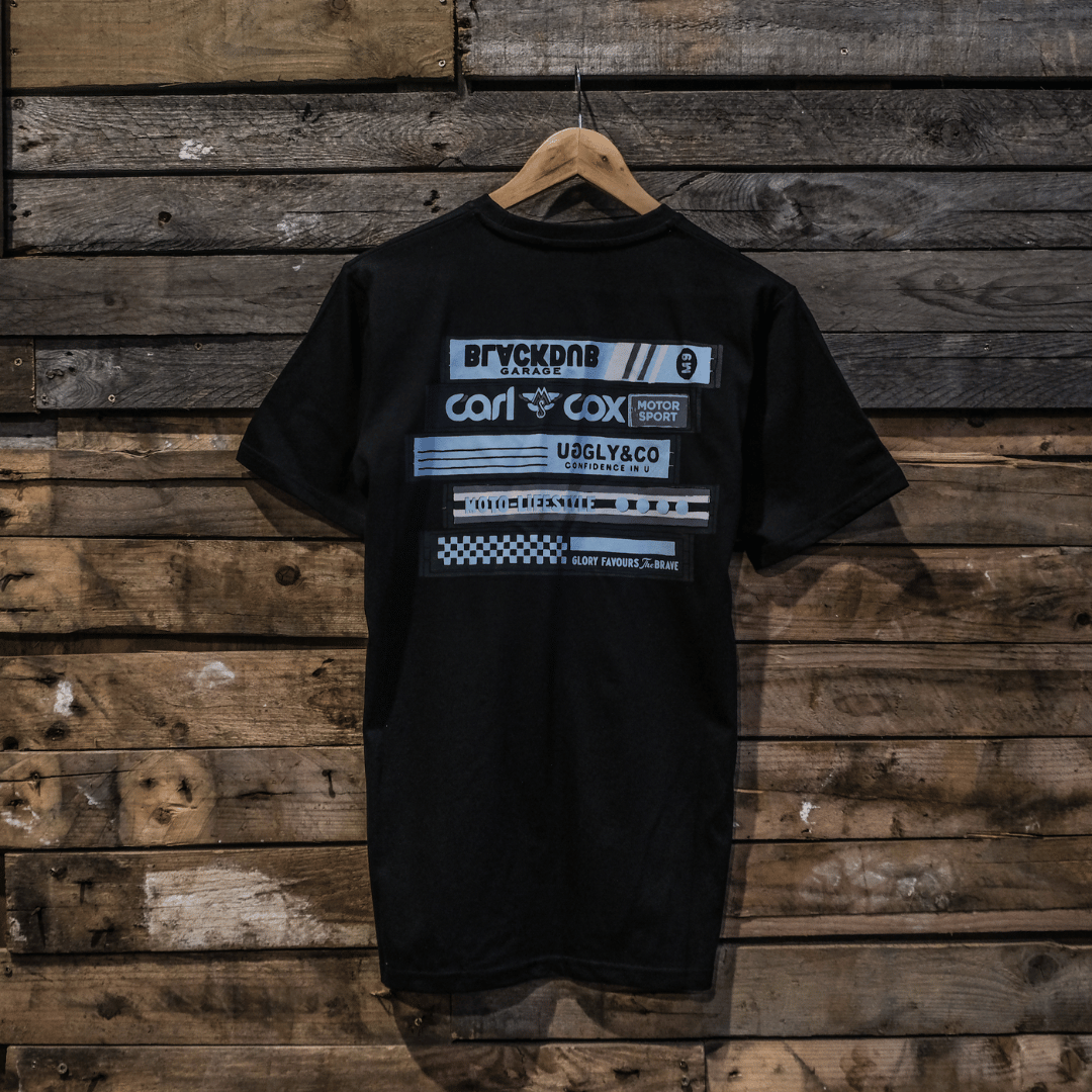 A black crew neck t-shirt with a graphic design in the center, featuring text and a cassette tape motif. The t-shirt is displayed on a wooden background.