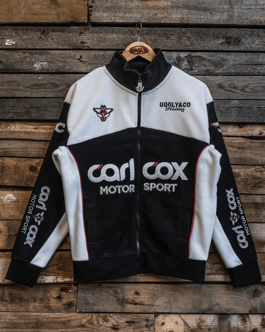A black and white Carl Cox Motorsport track jacket with embroidered logos and badges displayed on a wooden background.