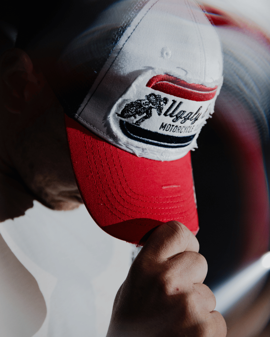 A model looking down while wearing a red, white and blue flat-track cap with a frayed Uggly&Co badge featuring a motorcycle racing theme with a blurred background
