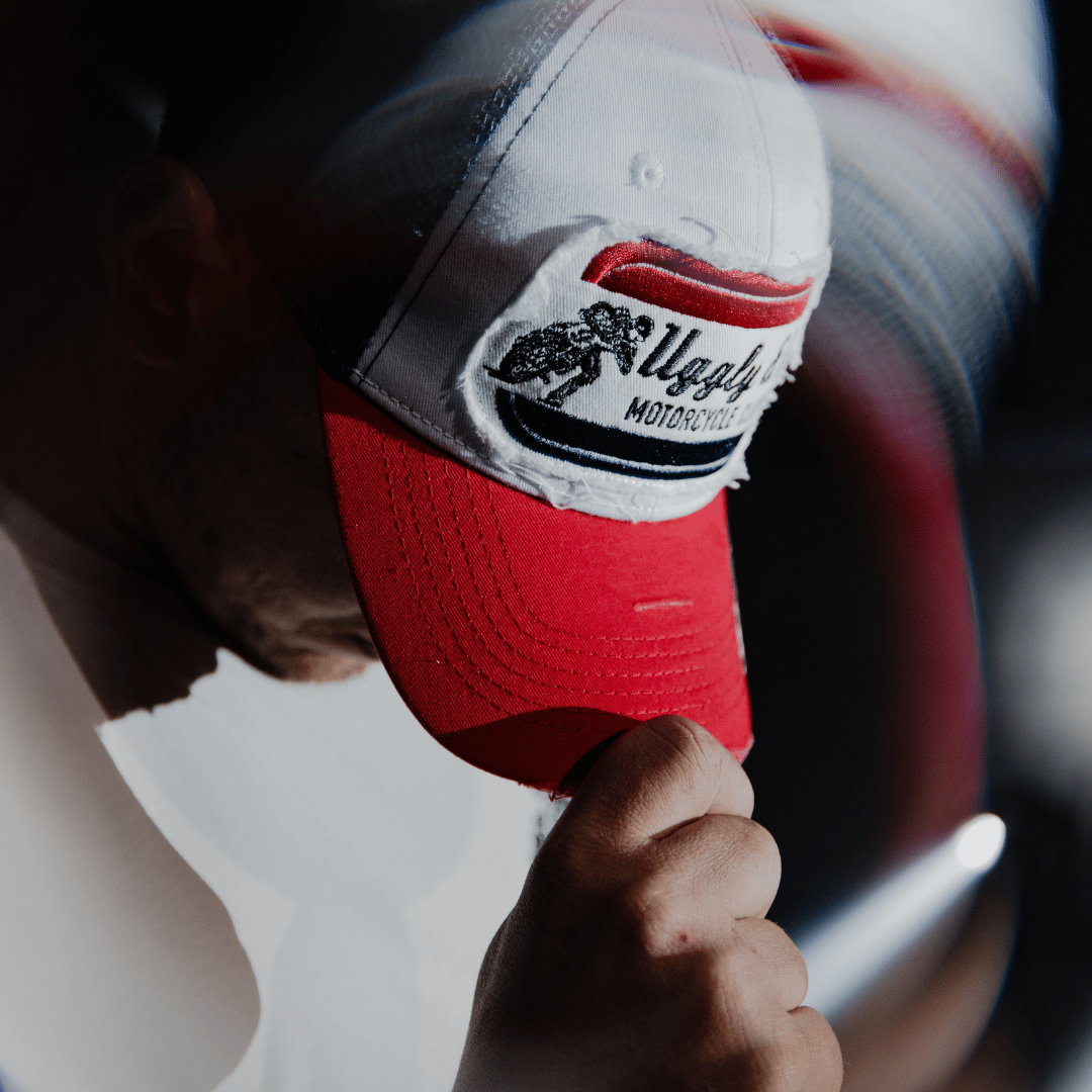 A model looking down while wearing a red, white and blue flat-track cap with a frayed Uggly&Co badge featuring a motorcycle racing theme with a blurred background
