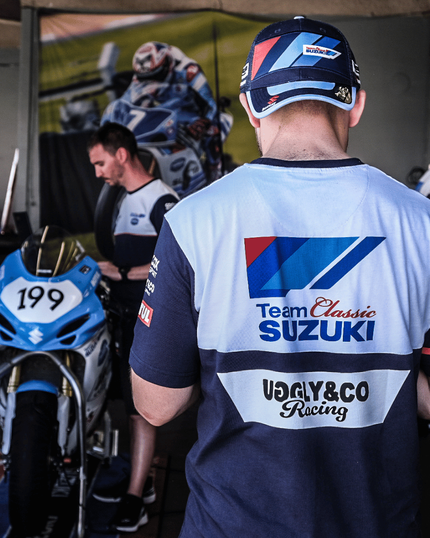 A person wearing a trucker hat backwards with geometric pattern featuring blue, red, and black colours, with the Team Classic Suzuki and Suzuki logos on it in the paddock