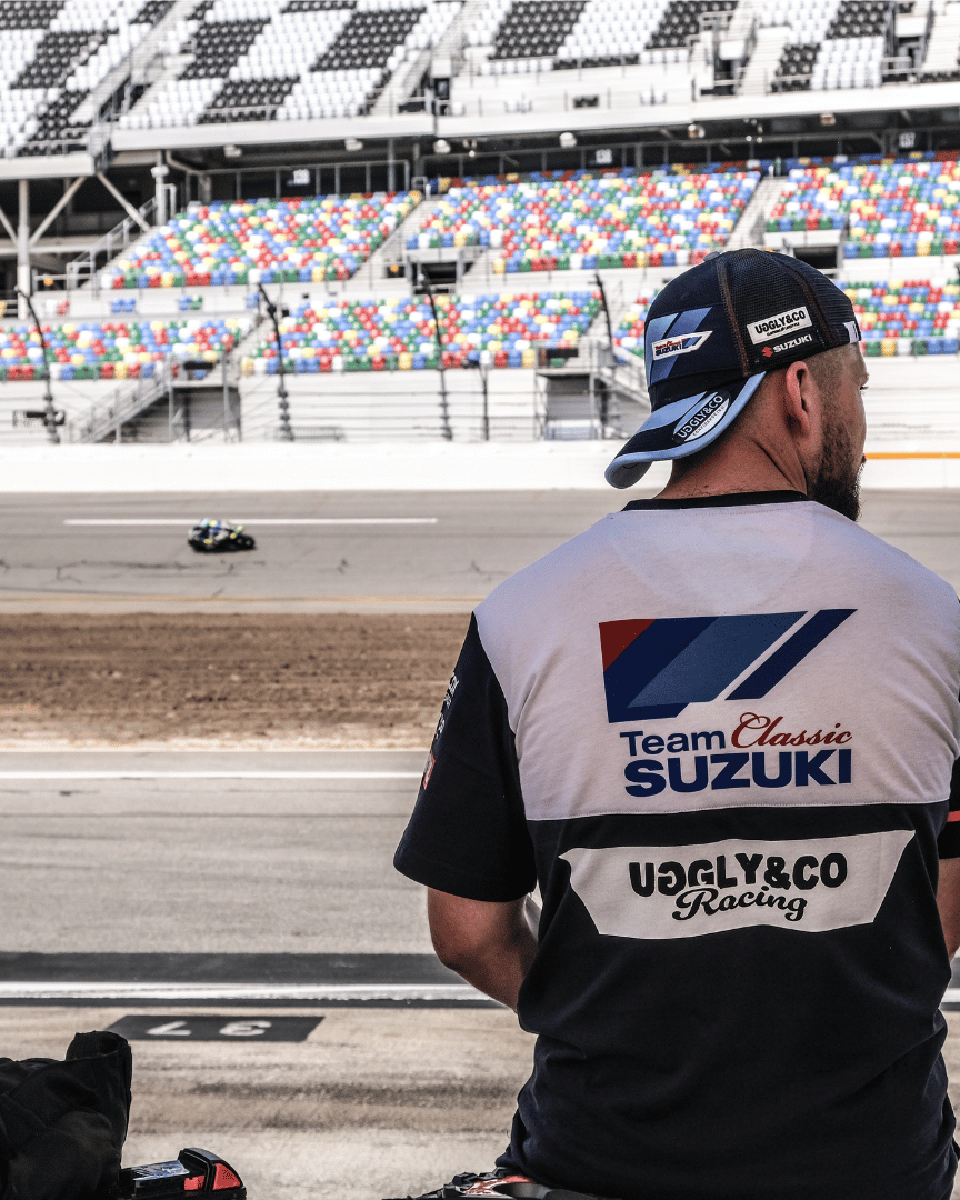 A model wearing a trucker hat backwards with geometric pattern featuring blue, red, and black colours, with the Team Classic Suzuki and Suzuki logos on it by a race track