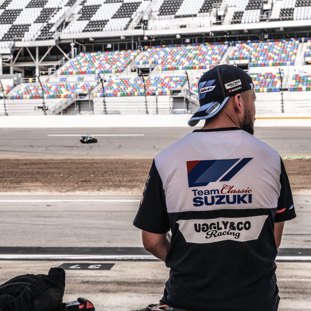 A model wearing a trucker hat backwards with geometric pattern featuring blue, red, and black colours, with the Team Classic Suzuki and Suzuki logos on it by a race track