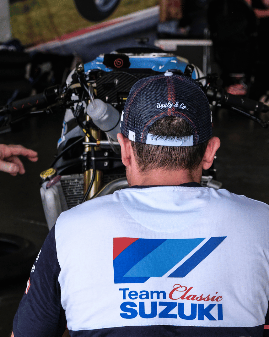 A model wearing a trucker hat backwards with geometric pattern featuring blue, red, and black colours, with the Team Classic Suzuki and Suzuki logos on it by a race track