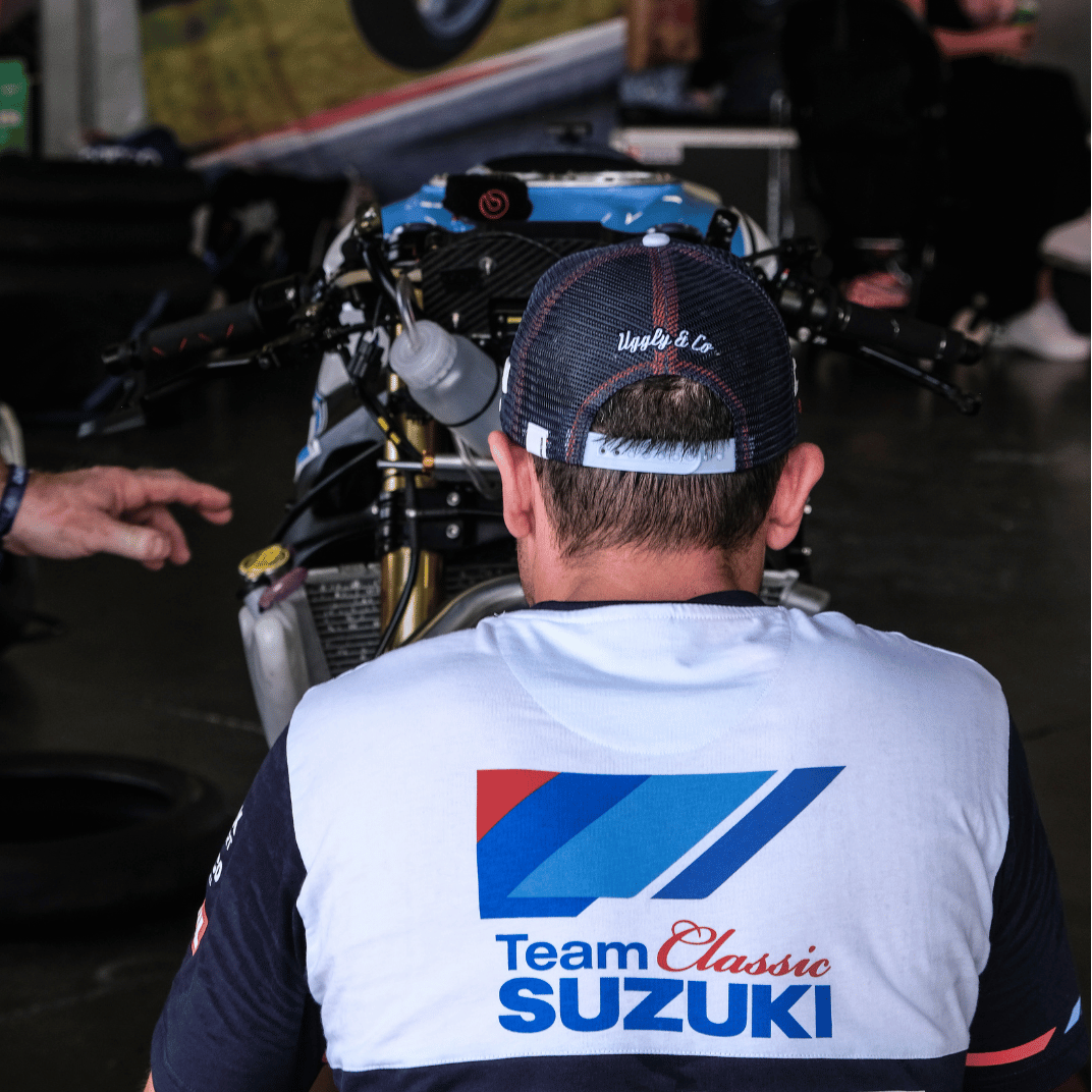A model wearing a trucker hat backwards with geometric pattern featuring blue, red, and black colours, with the Team Classic Suzuki and Suzuki logos on it by a race track
