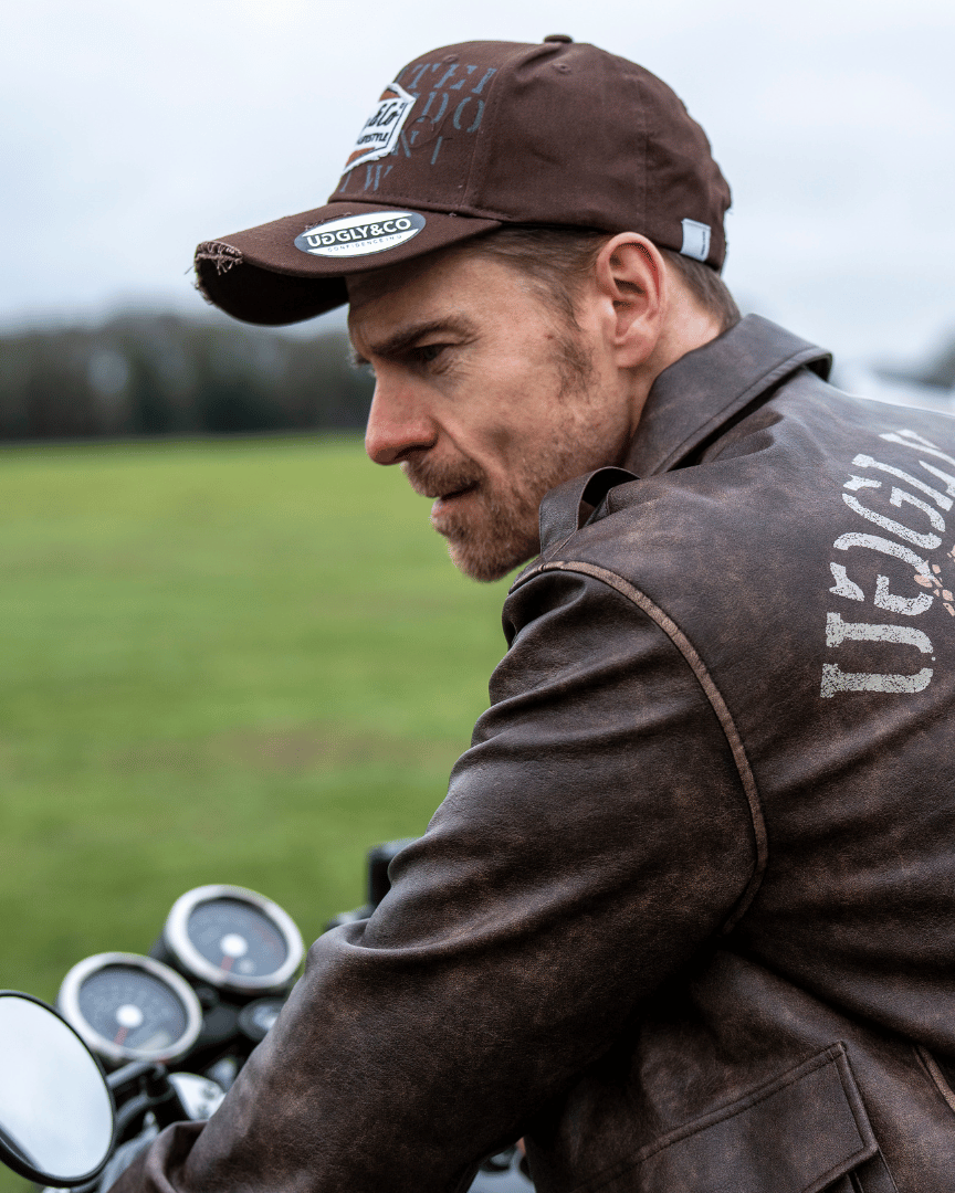 Side view of a man wearing a brown 'Aviation' Diamond Trucker Hat with the Uggly&Co logo on the front and brown leather jacket sitting on a motorcycle