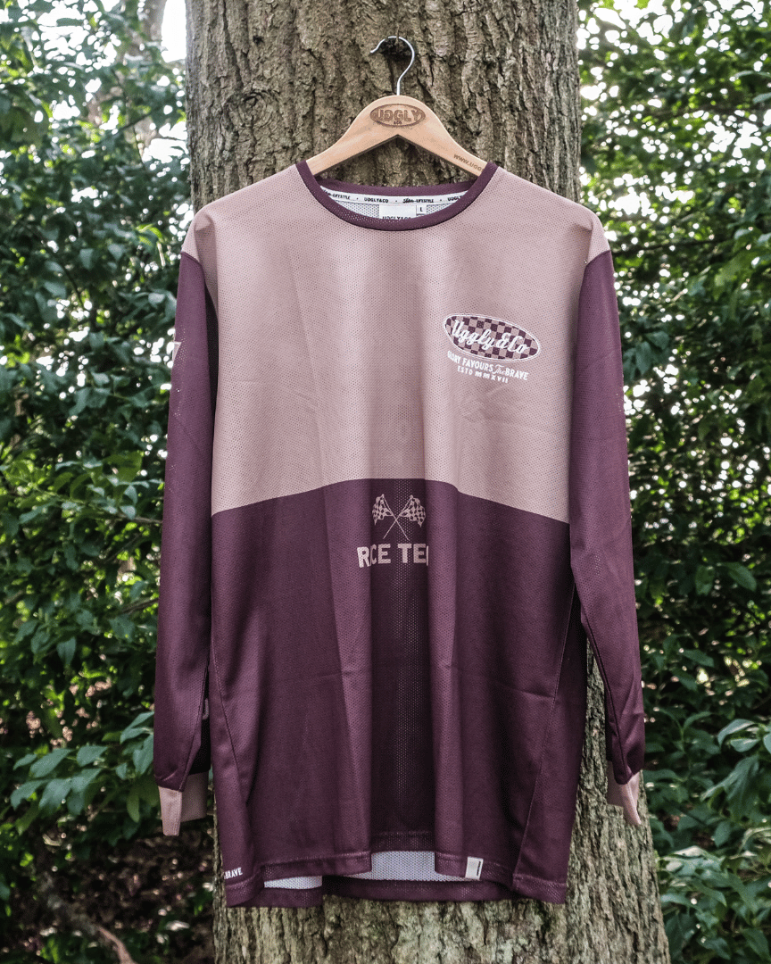 A long-sleeved brown and beige motocross jersey with checkered patterns and text graphics hanging on a tree forwards.