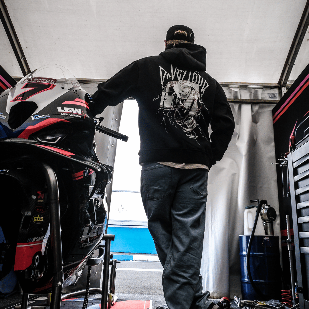 Davey Todd wearing a black hoodie with a graphic design, standing next to a motorcycle in a garage.