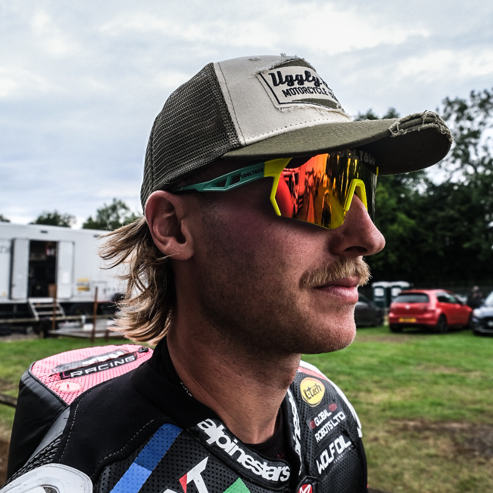 Close up view of Davey Todd wearing a green and beige trucker hat with a distressed design and an Uggly&Co logo on the front, with trees in the background