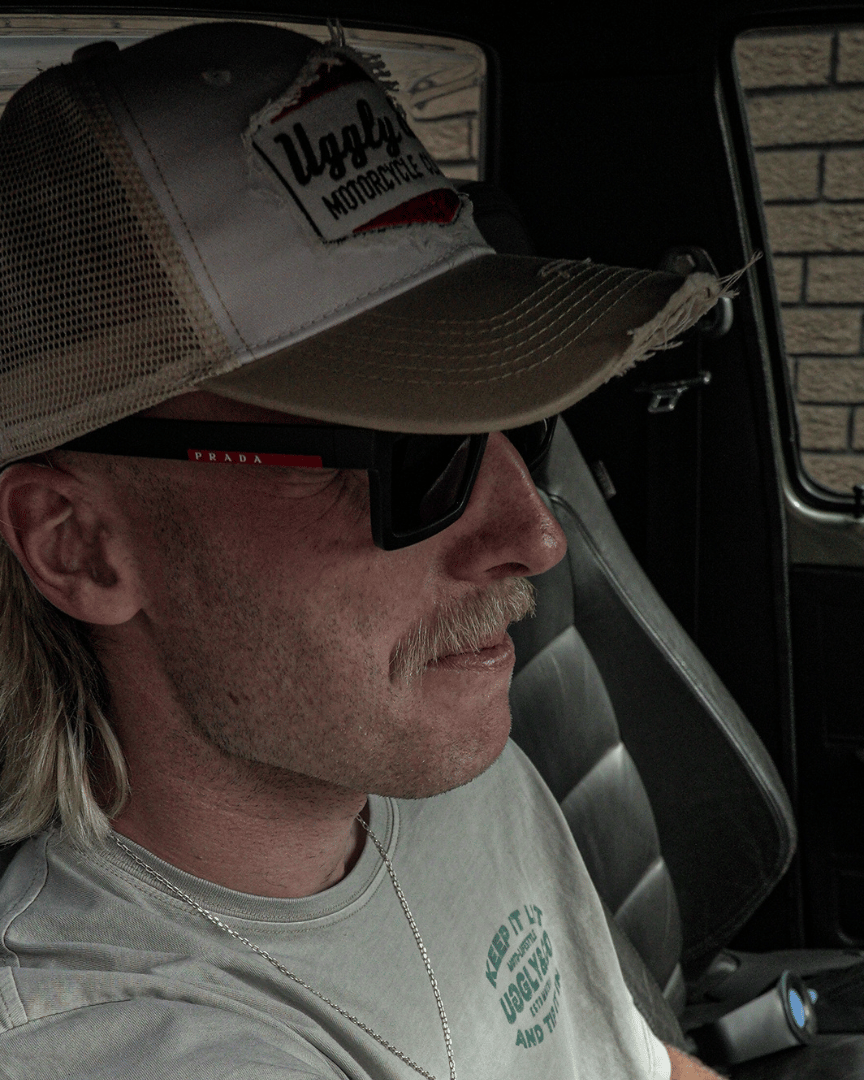 A model sitting in a car driver's seat while wearing a beige and white Uggly & Co motorcycle clothing trucker hat with a mesh back and an embroidered logo on the front
