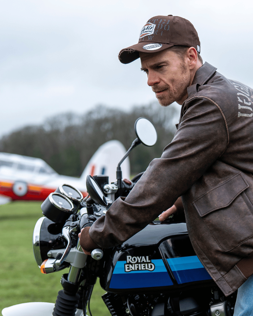 A man wearing a brown jacket with 'Uggly & Co' branding, sitting on a motorcycle on a grassy field.