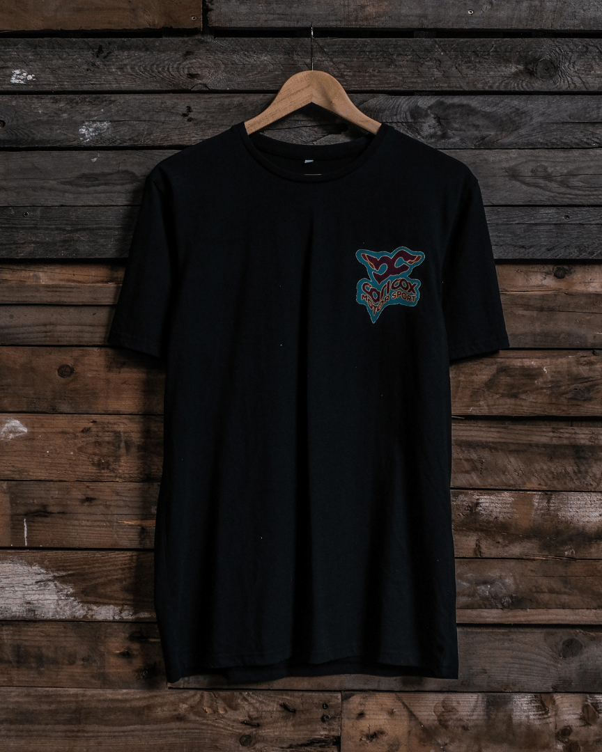 Front view of a Carl Cox motorsport black t-shirt with a colourful graphic design on a wooden wall