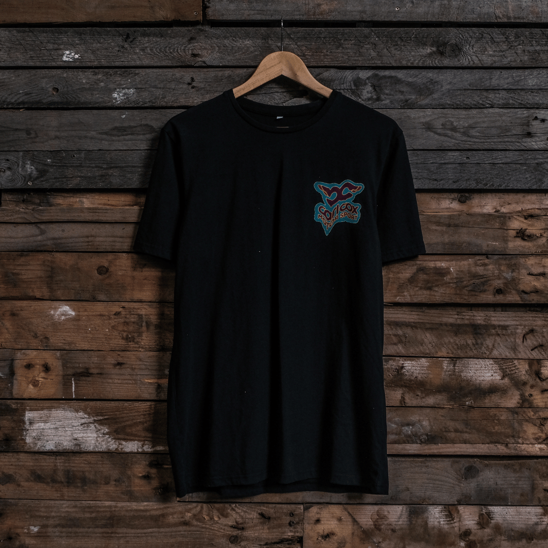 Front view of a Carl Cox motorsport black t-shirt with a colourful graphic design on a wooden wall