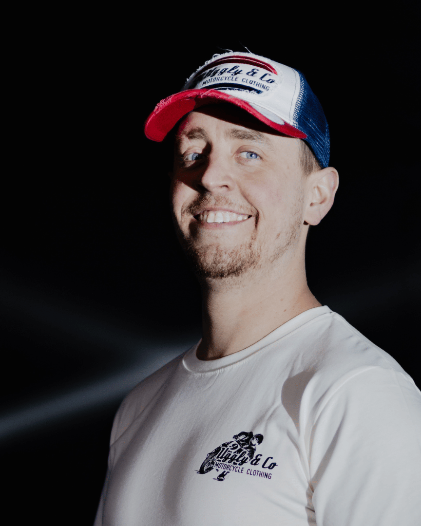 A model looking at the camera and smiling while wearing a red, white and blue flat-track cap with a frayed Uggly&Co badge featuring a motorcycle racing theme