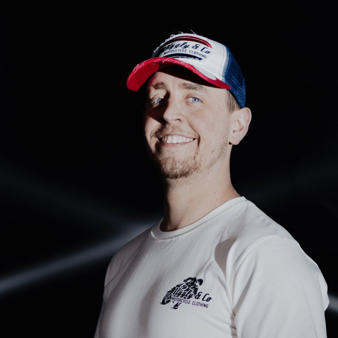 A model looking at the camera and smiling while wearing a red, white and blue flat-track cap with a frayed Uggly&Co badge featuring a motorcycle racing theme