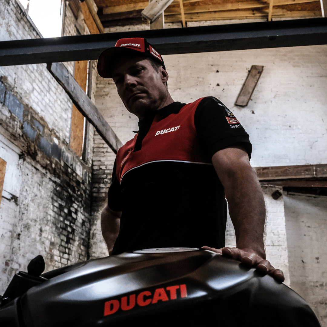 A model looking down at a motorcycle while wearing a Milwaukee x Ducati polo shirt, featuring a black lower half and a red upper torso in a warehouse