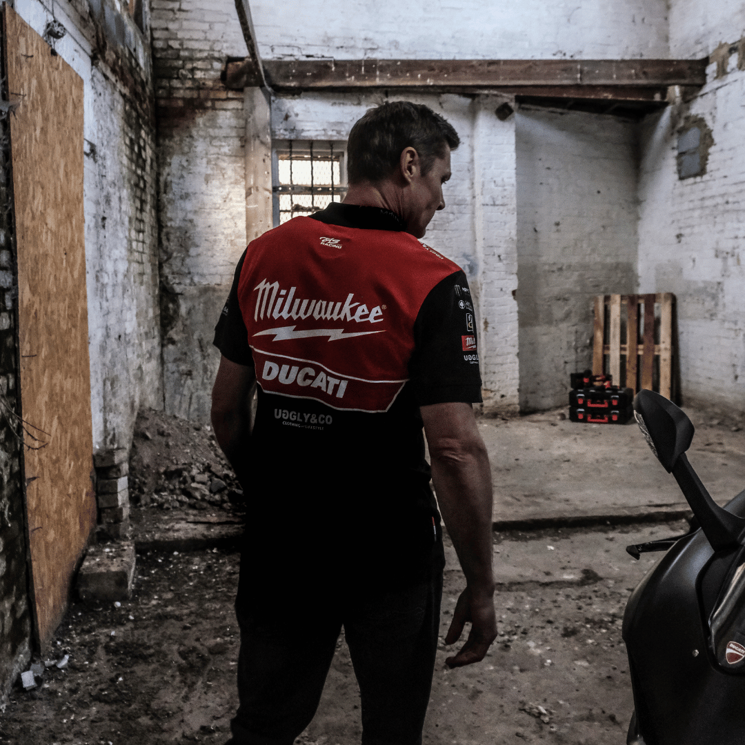 A model walking away and looking right while wearing a Milwaukee x Ducati polo shirt, featuring a black lower half and a red upper torso in a warehouse