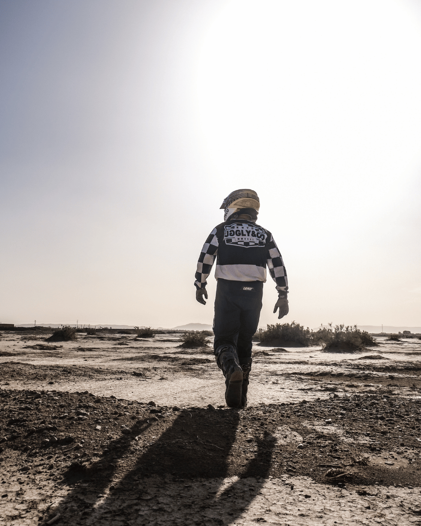 Motocross rider in full gear in the desert wearing a chequered Uggly&Co MX shirt walking away from the camera