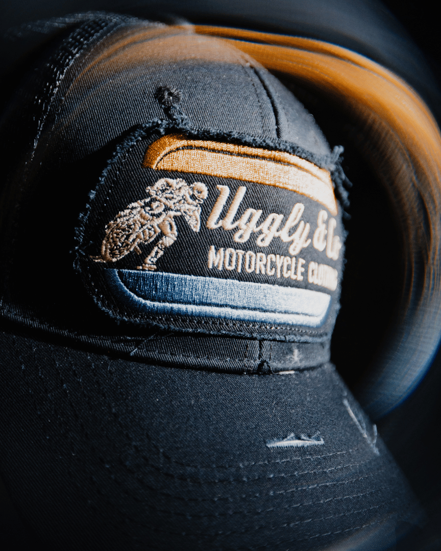 An all-black trucker hat with a vintage-inspired flat track racing logo in yellow and blue on the front at night with a spinning blurred background