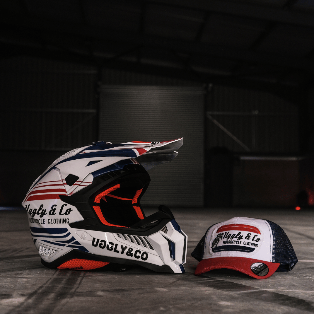 A red, white and blue flat-track cap with a frayed Uggly&Co badge featuring a motorcycle racing theme, on the floor with a matching motocross helmet with a blurred garage background