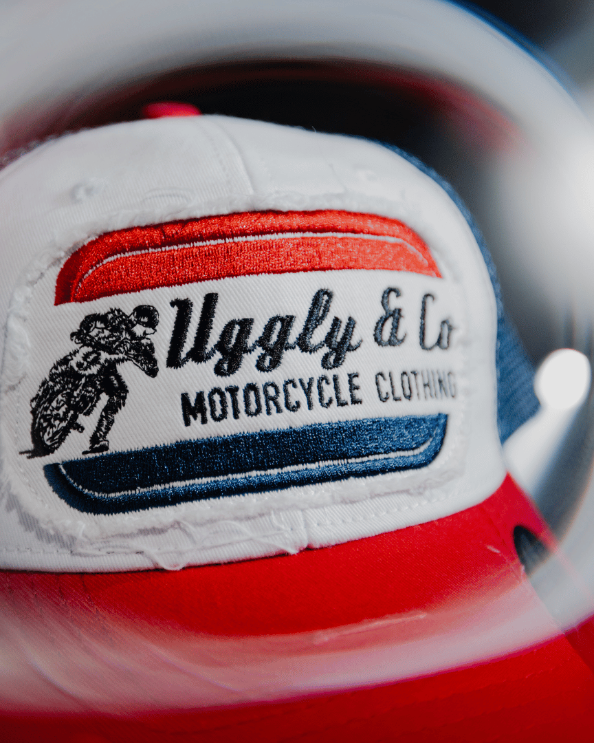A close up of the front of a red, white and blue flat-track cap with a frayed Uggly&Co badge featuring a motorcycle racing theme